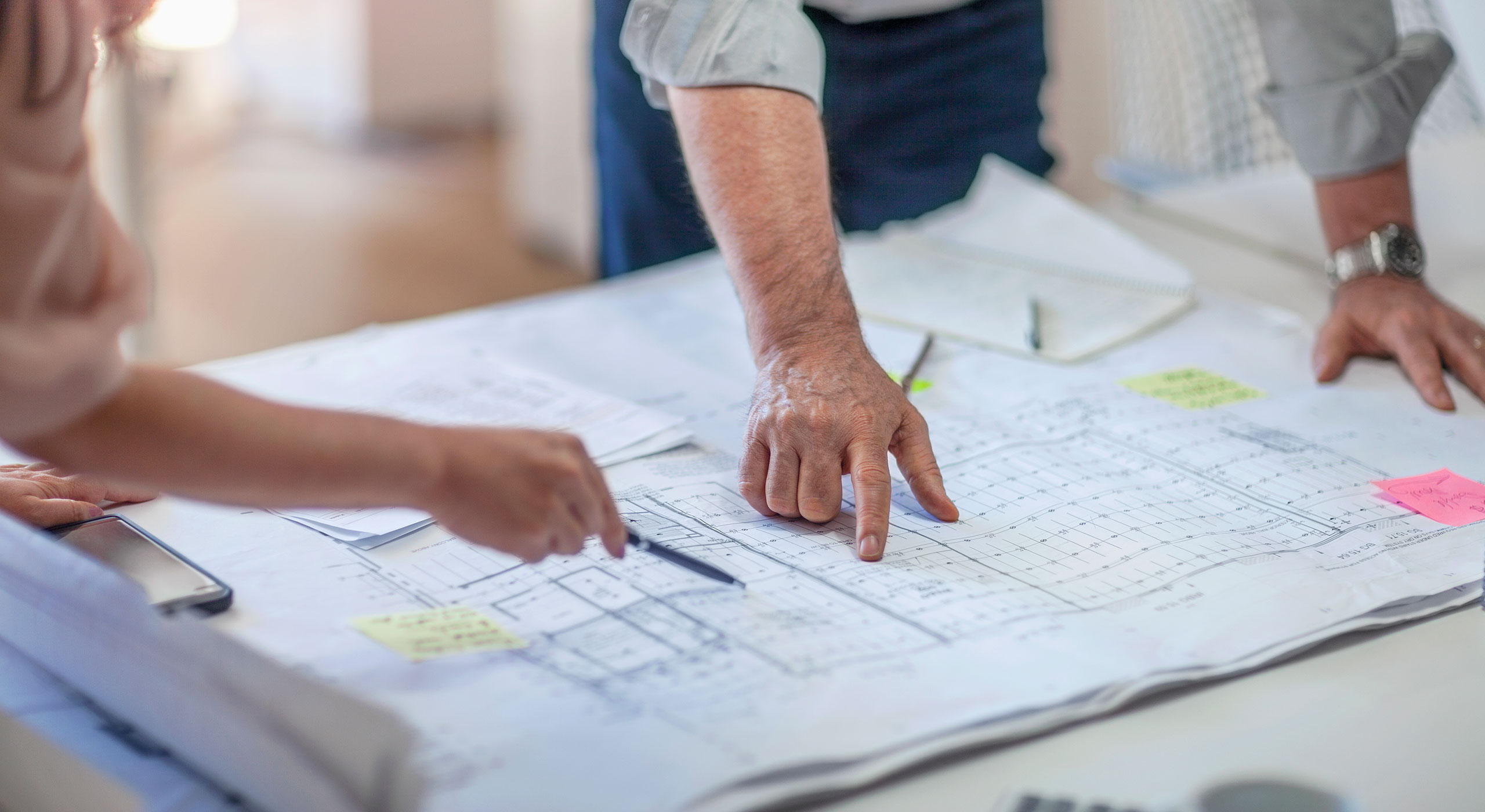 Hands of professionals pointing at building blueprints during a planning session, illustrating Woolpert’s program management services for complex projects.
